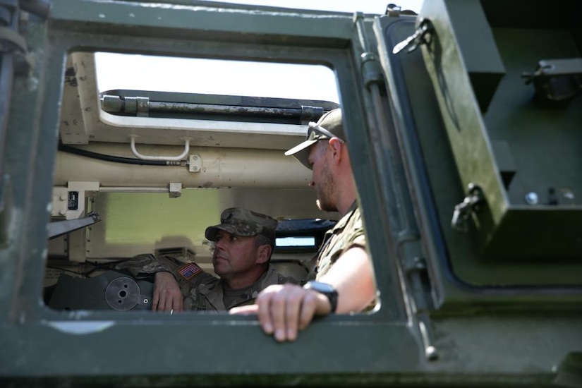 A uniformed service member explains the capabilities to another service member while inside of a tank.
