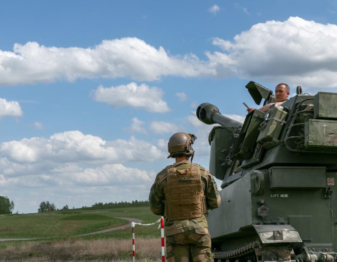 A service member sitting in the top of a tank looks at another uniformed service member who is standing on the ground.