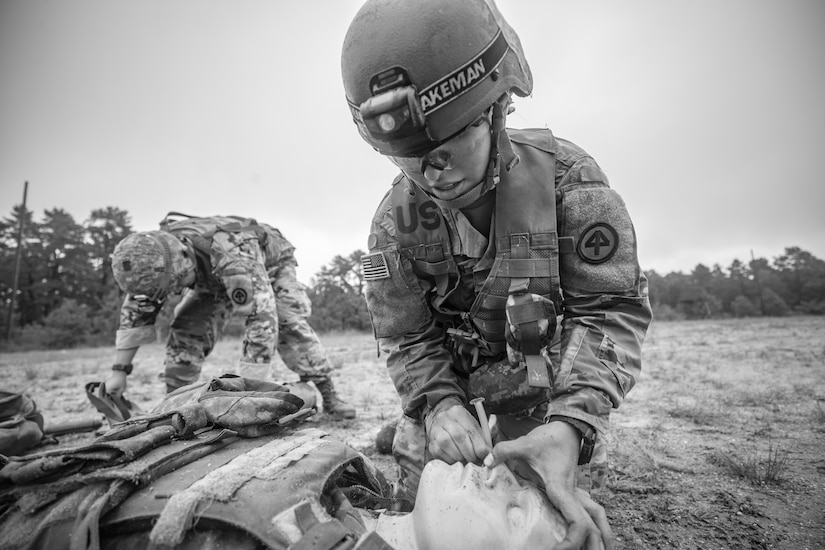 A uniformed service member kneels over a training mannequin in a field while another uniformed service member sifts through gear in the background.