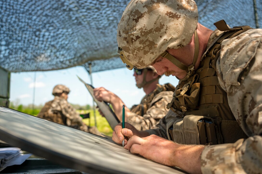 A Marines plots points on a board under a tent as fellow service members work nearby.