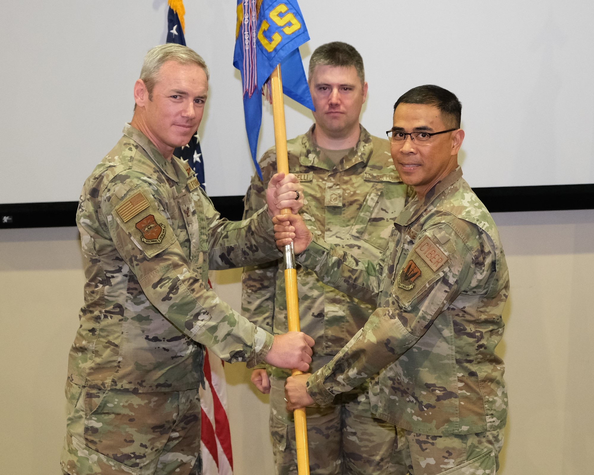 Alt caption: photo of three U.S. Air Force Airmen, two standing holding a unit guidon with Airmen standing in the background.
