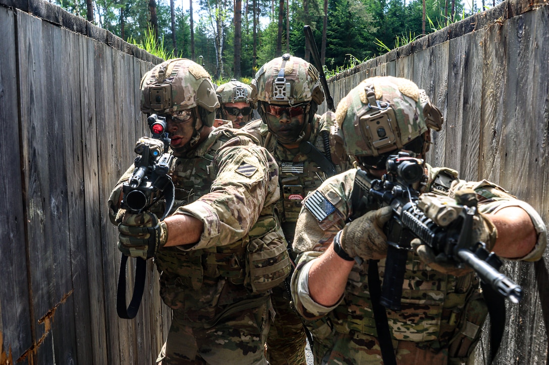 Four soldiers with camouflage face paint and weapons move through a narrow path with wood walls in a forest area.