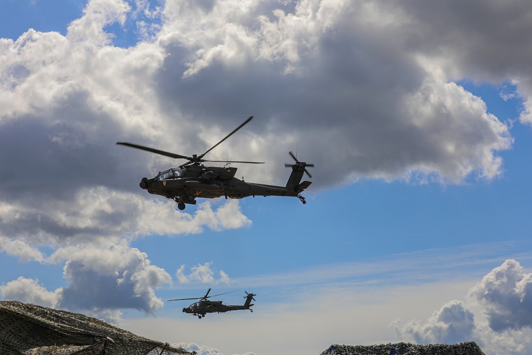 Two military helicopters fly in a partly clouded sky.