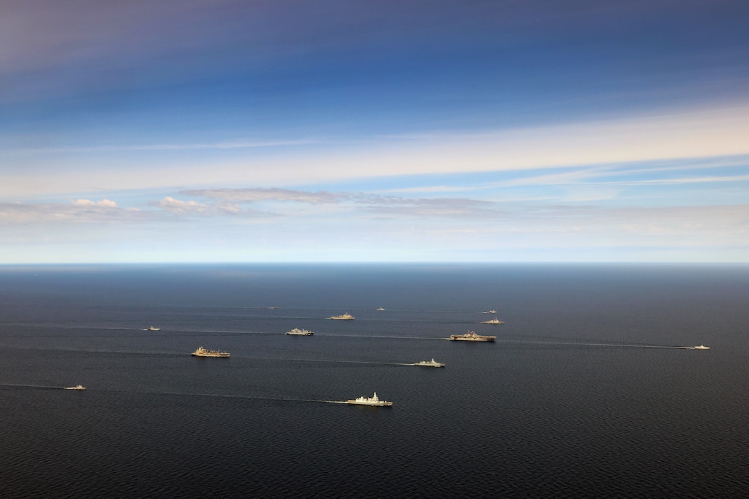 Thirteen naval ships sail in formation in a sea.