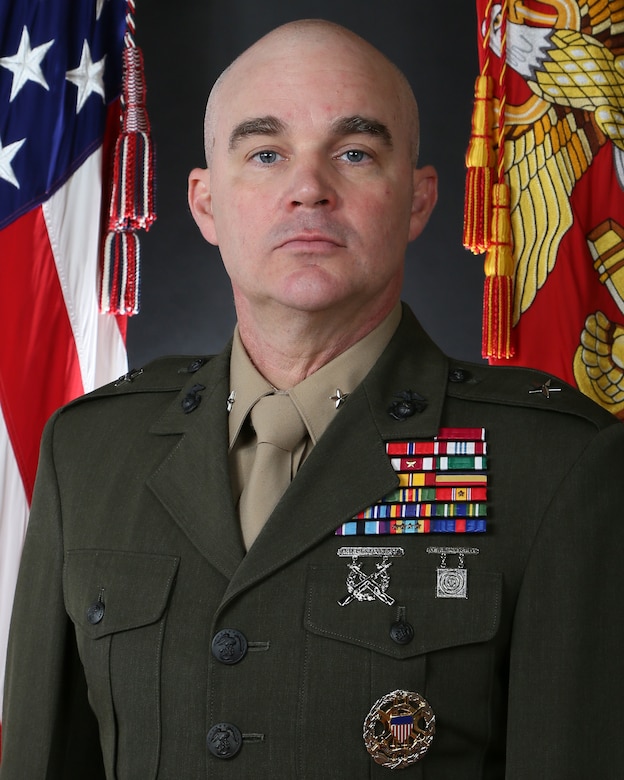 A headshot of a Marine in uniform with the American flag and Marine Corps flag in the background.