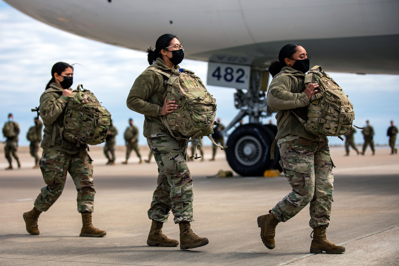 Three female soldiers wearing face masks carry backpacks across the tarmac. A large airplane is in the background. A long line of soldiers stretches behind them.