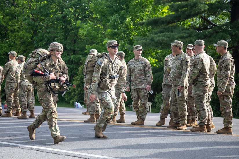 Two soldiers wearing large backpacks walk on a road past a large group of soldiers.