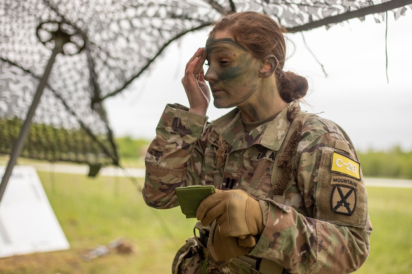 A female soldier rubs green camouflage paint onto her face.