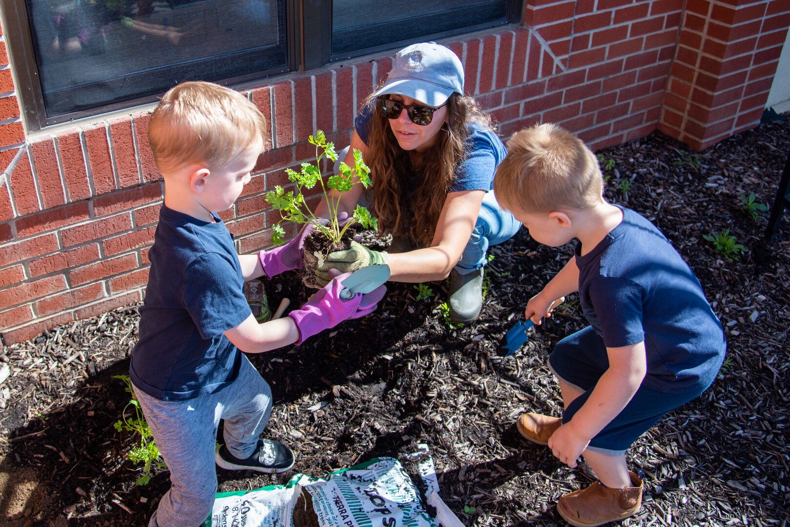 A woman in jeans, a baseball cap and sunglasses teaches children how to plant a cilantro plant in the CDC garden.