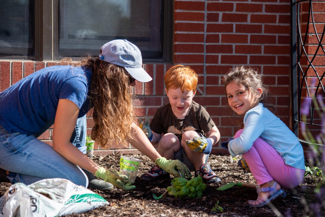 A woman in a baseball cap helps two children plant a plant in the CDC garden.