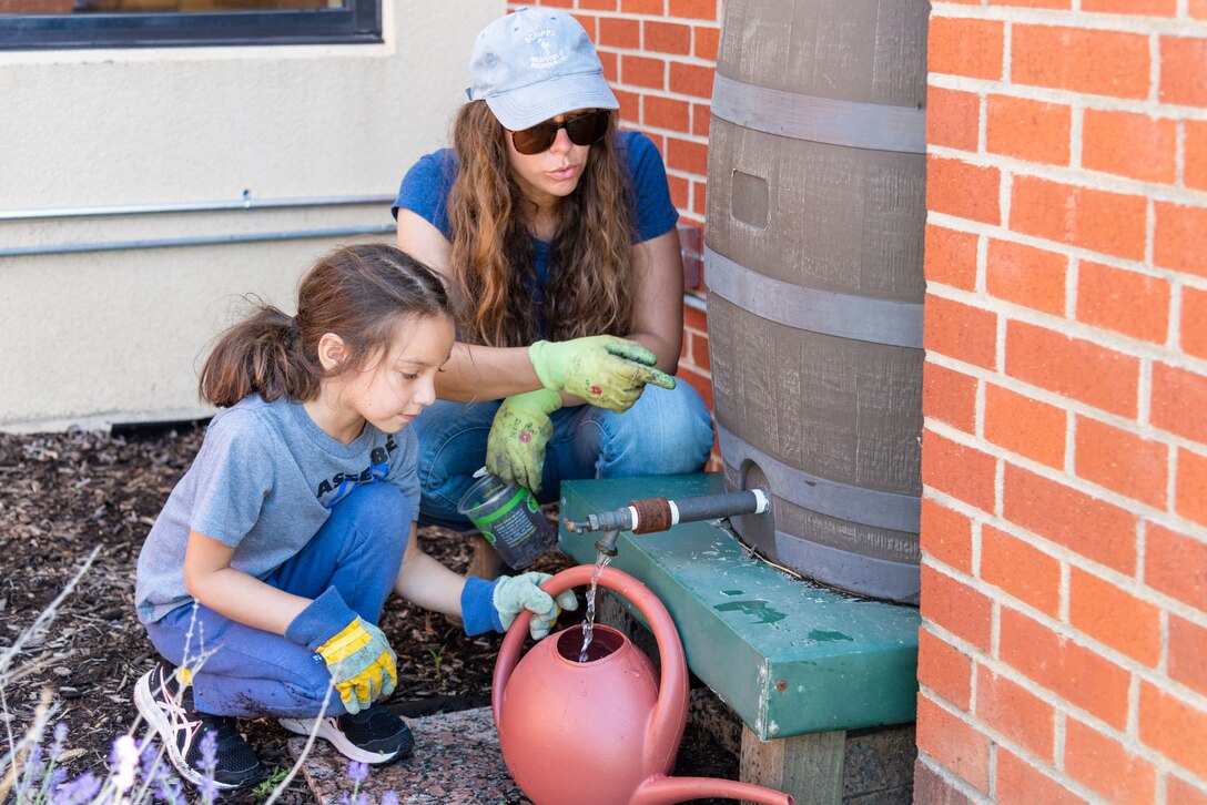 A woman in a baseball cap and jeans  shows a child how to get water from a rain barrel.