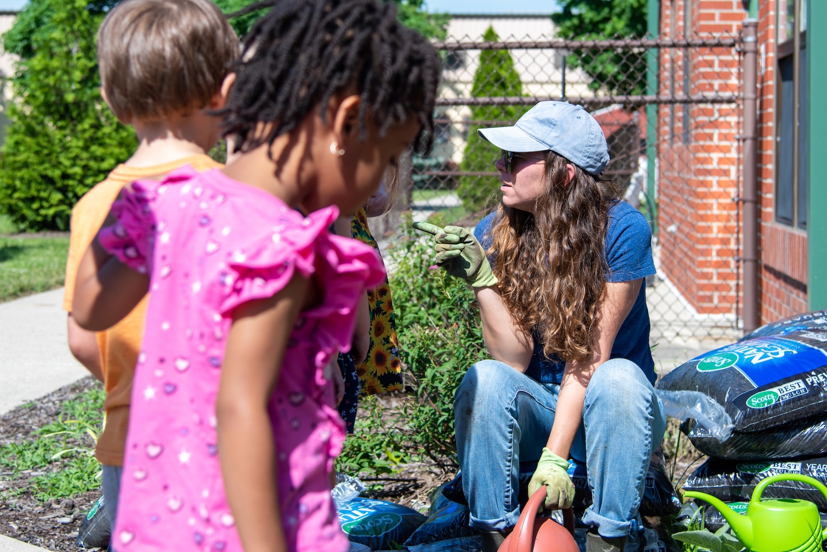 A woman in jeans in a baseball cap and sunglasses talks to a grouip of children.