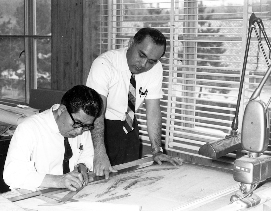 Man drawing at desk while another man looks over his shoulder.