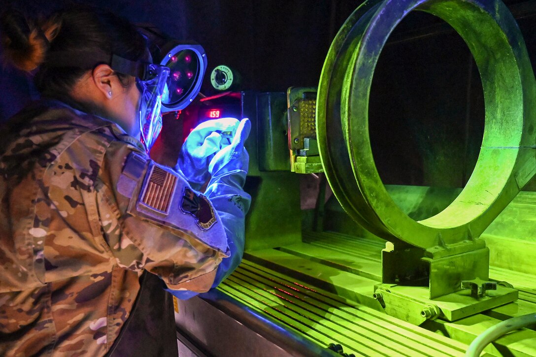 An airman wearing a face shield looks a small objects near large machinery illuminated by green and blue lights.