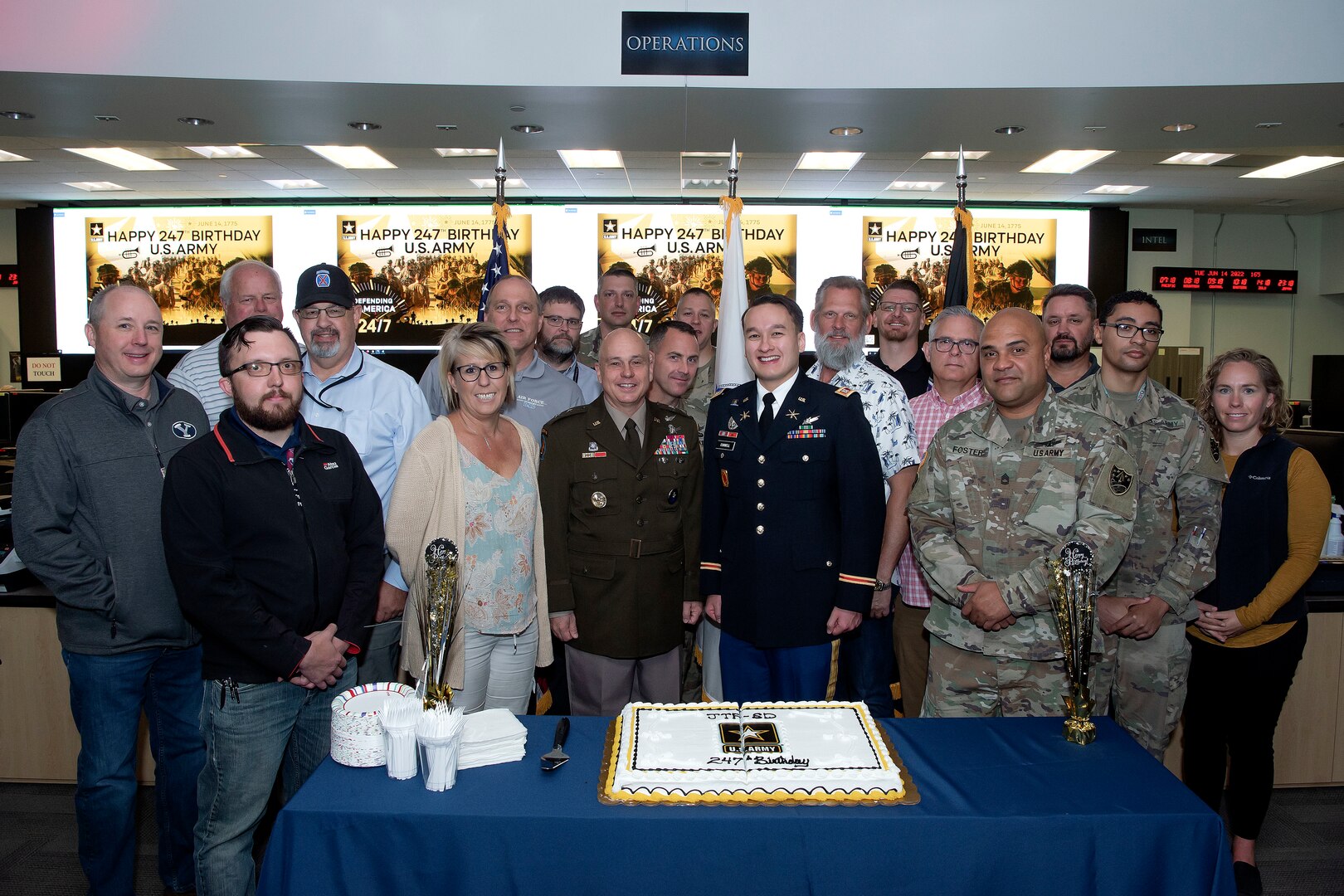 Group photo of many people standing in front of table with cake smiling