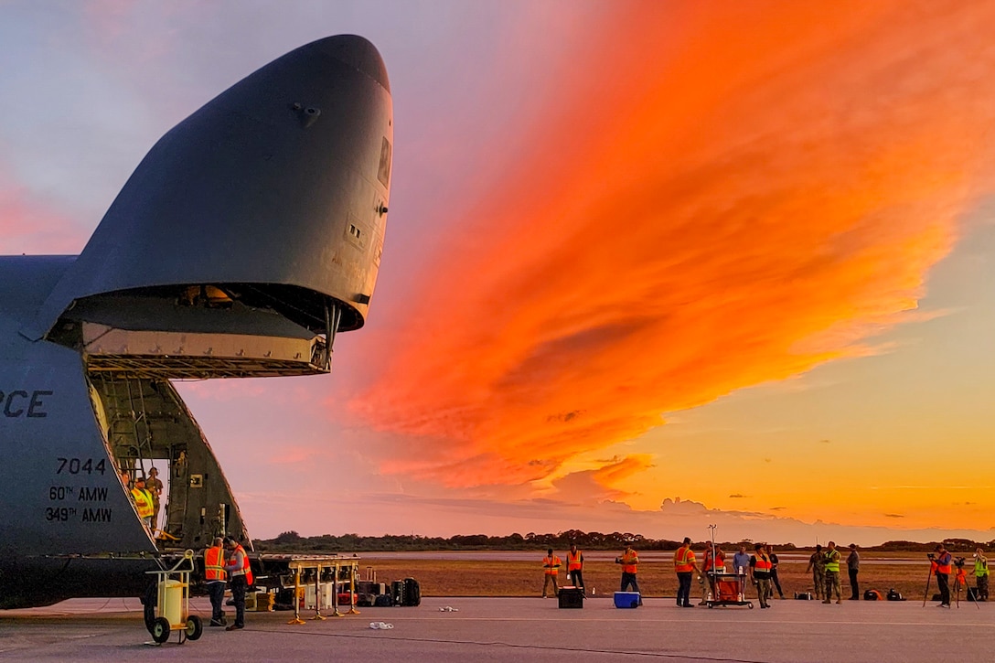Airmen unload a satellite from an aircraft under a sunlit sky.