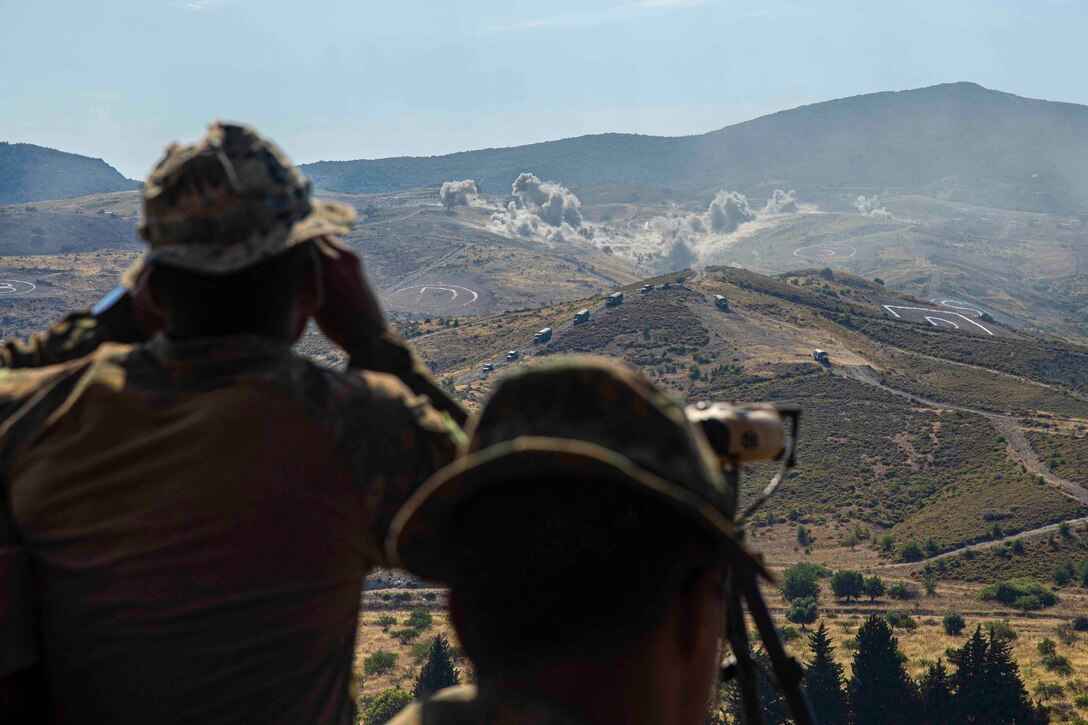 Two Marines observe artillery fire on a mountain range.