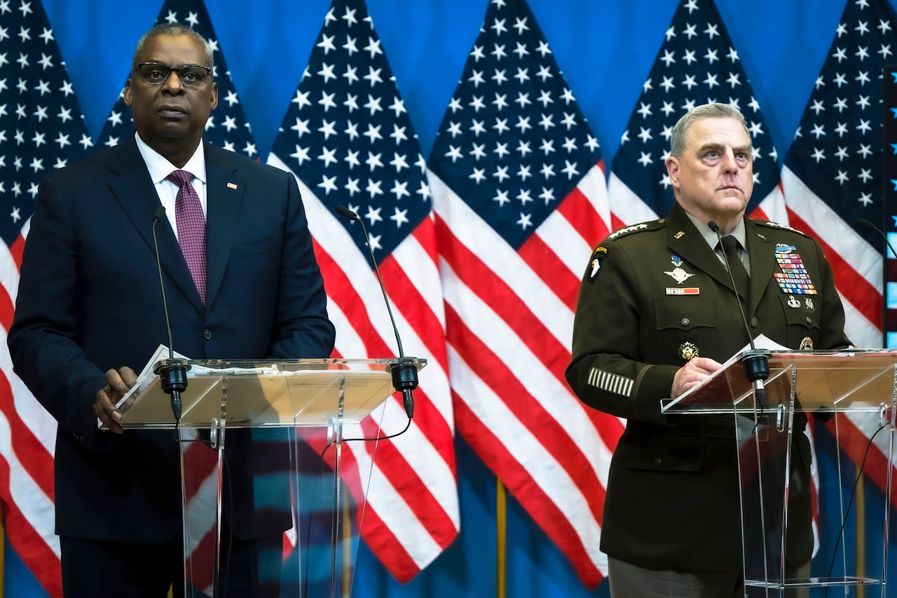 Two men stand on a stage with flags behind them.