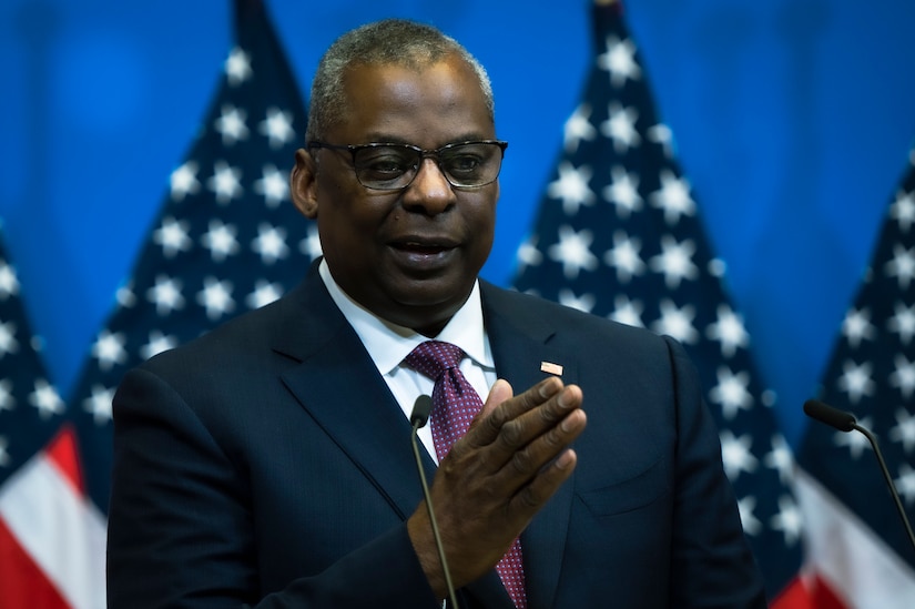 A man wearing a business suit speaks into a microphone with several American flags in the background.