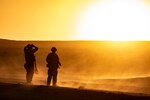 Two soldiers stand in a desert environment, silhouetted by the sun.