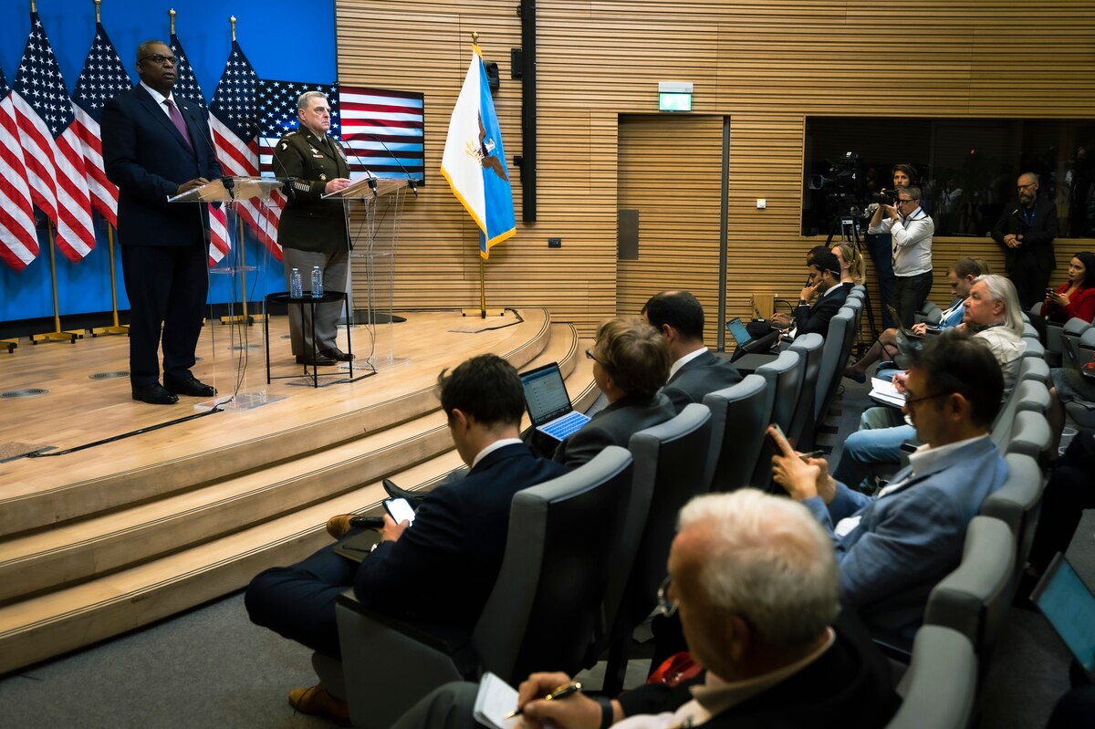 Two people, one person in a military uniform, stand on stage behind two separate lecterns in front of a crowd.