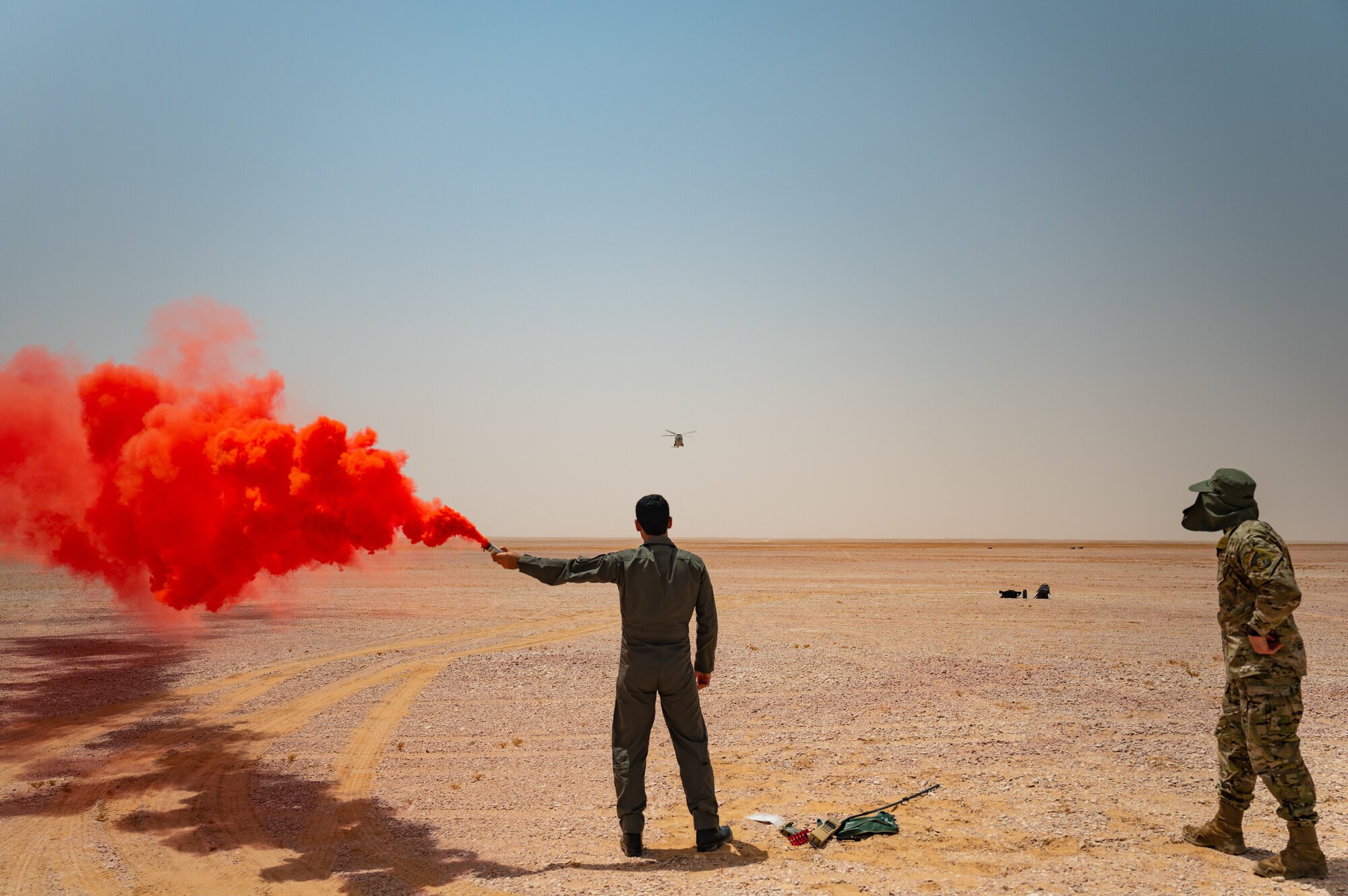 A Royal Air Force of Oman pilot, center, uses a smoke flare to indicate his position of retrieval to a rescue helicopter with U.S. Air Force Tech. Sgt. David Jones, right, a survival evasion resistance and escape instructor from U.S. Air Forces Central A3 Operations, while performing a survival training scenario in the Oman desert during Accurate Test 22, May 17, 2022. This joint force training helped streamline processes and provided both forces a better understanding of mutually beneficial combat search and rescue operations. (U.S. Air Force photo by Staff Sgt. Dana Tourtellotte)