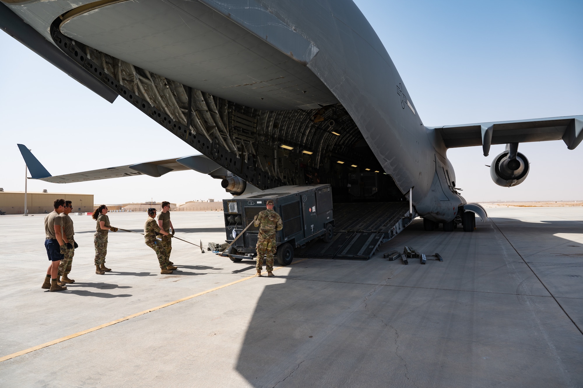U.S. Air Force Airmen from the 379th Air Expeditionary Wing load a mobile air conditioning unit onto a U.S. Air Force C-17 Globemaster in preparation for a departure flight during exercise Accurate Test 22 at Thumrait Air Base, Oman, May 22, 2022. The Airmen pictured steered the air conditioning unit from the back as a winch inside of the aircraft pulled the cargo up the ramp and into the plane. (U.S. Air Force photo by Staff Sgt. Dana Tourtellotte)