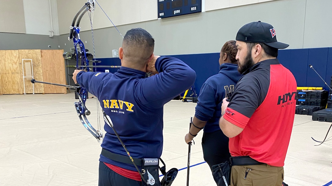 An athlete practices archery with assistance from a coach in a gym.