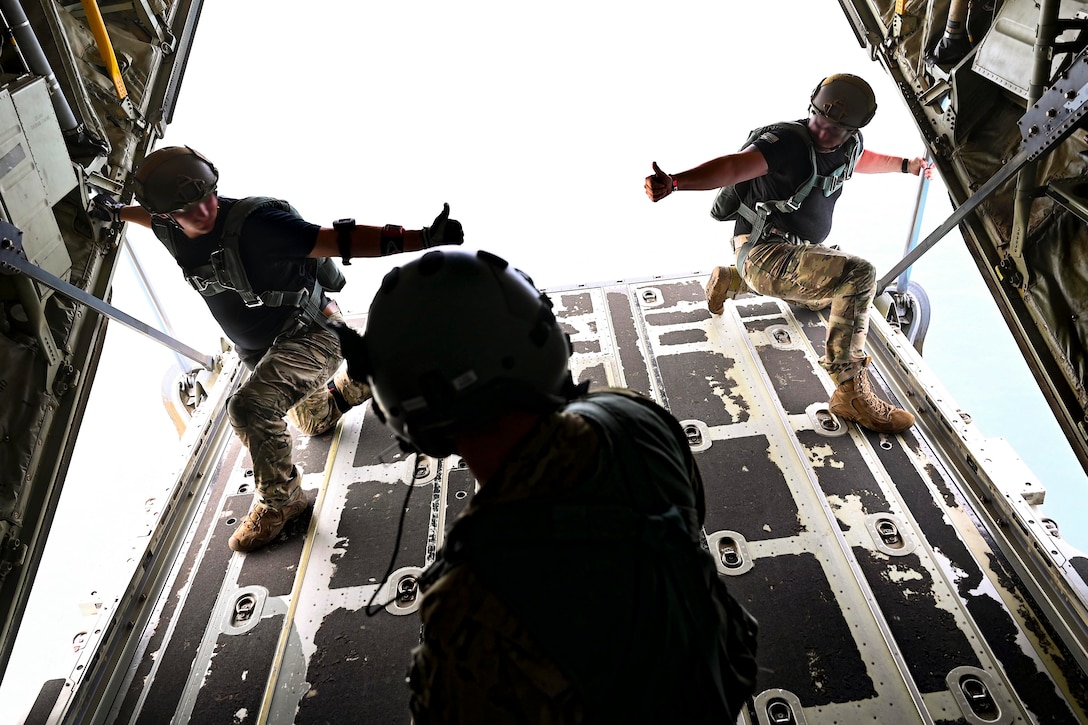 Soldiers give each other a thumbs up while standing on an open door of an airborne aircraft.