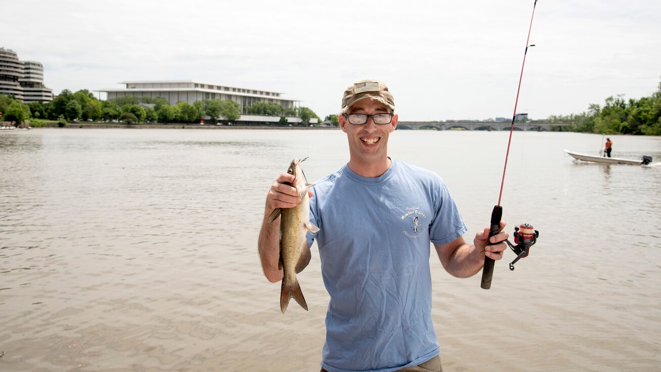 A participant holds up a catch fish he caught in the Potomac River with the Kennedy Center in the background.