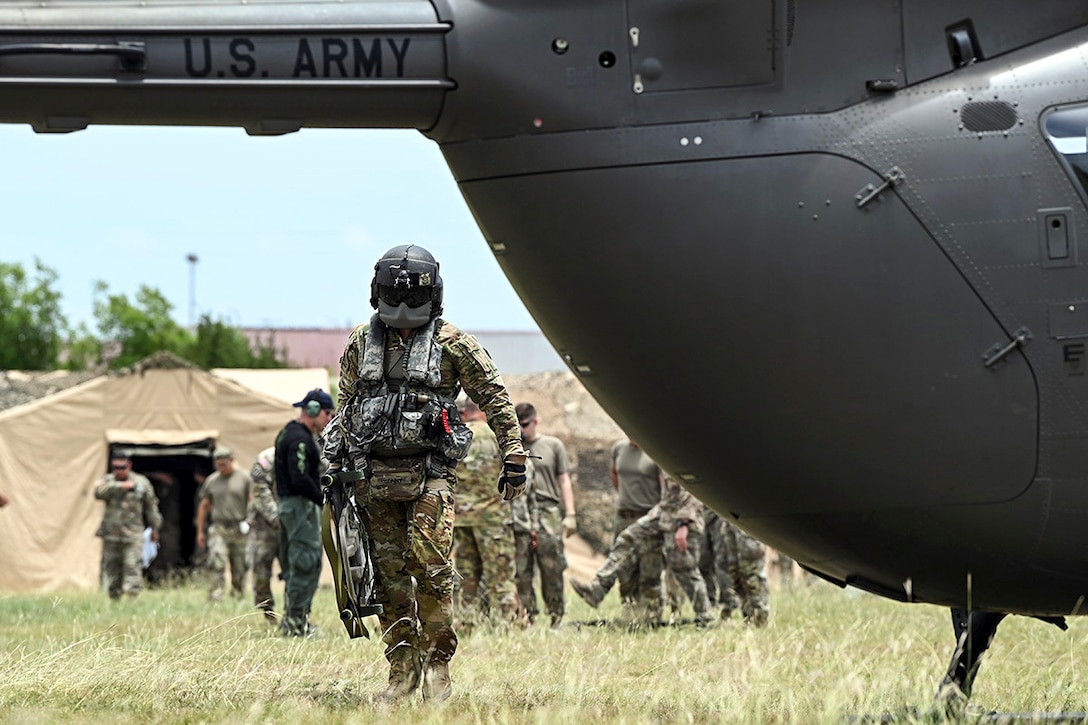 A soldier wearing a helmet and carrying a litter walks toward a helicopter parked in a field.