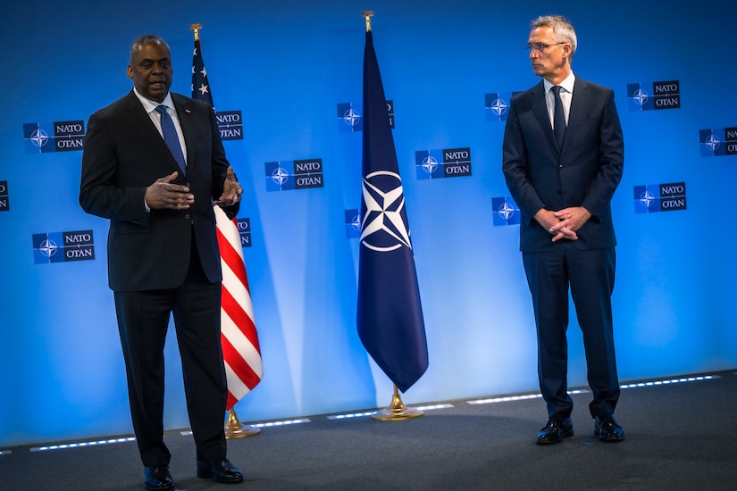 Two men wearing business suits stand on a stage with two flags in the background.