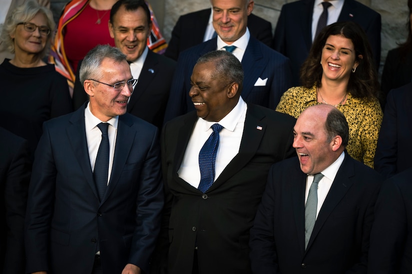 Three men wearing business share a humorous moment while several others in the background smile and look on.