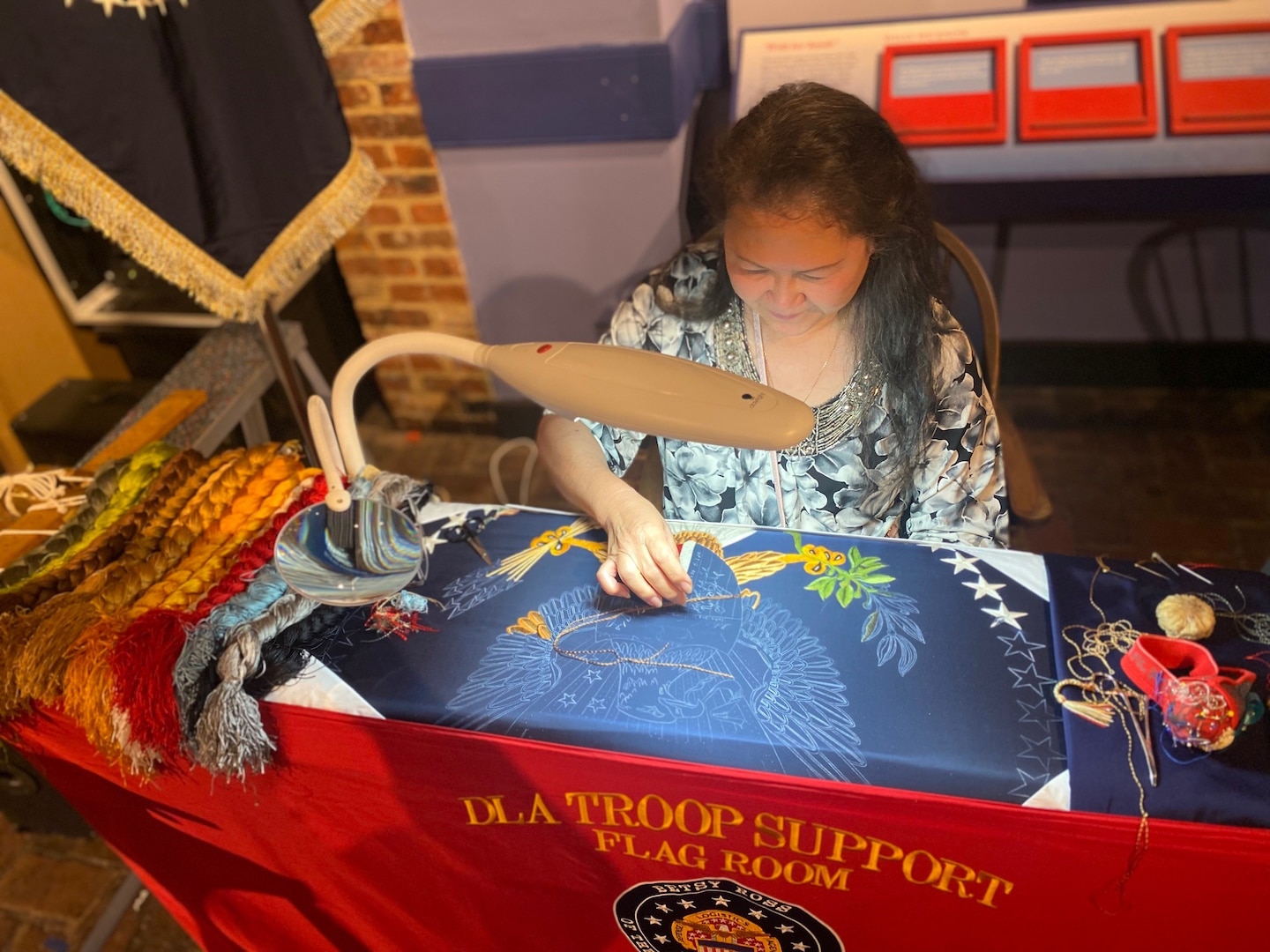 man and woman stand next to woman embroidering flag