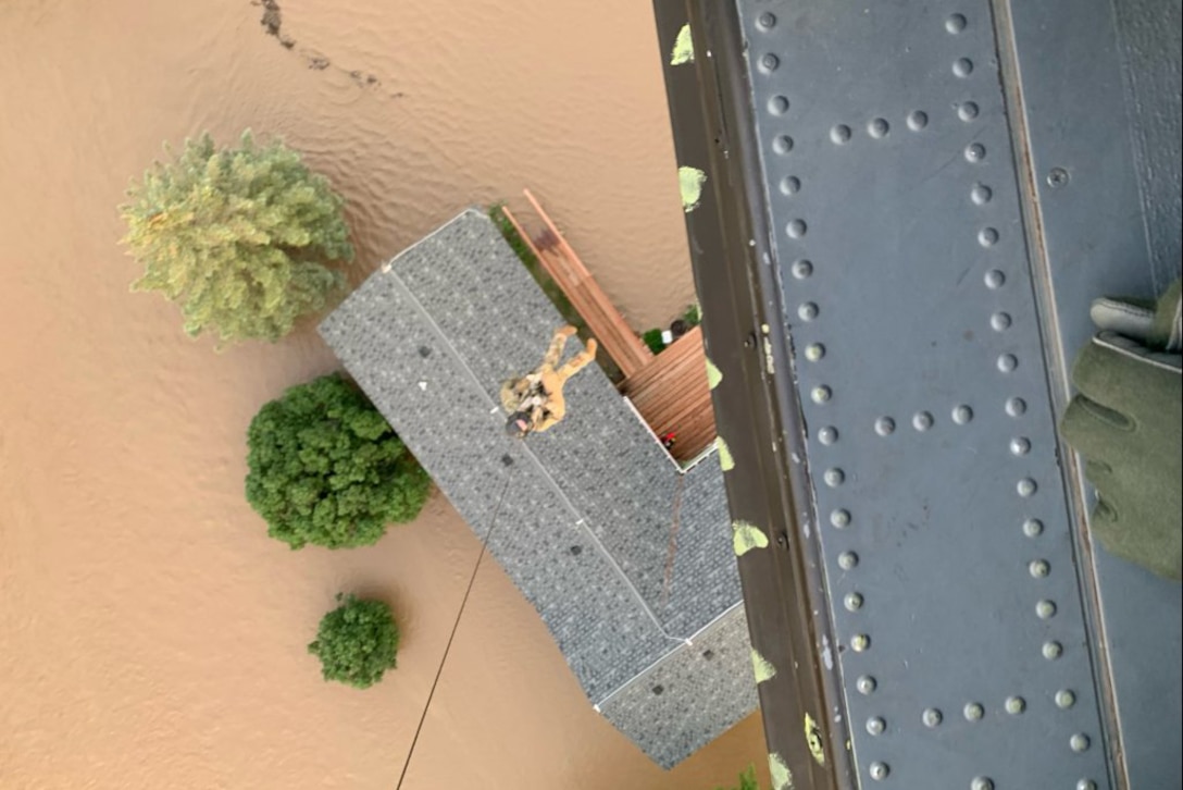 A National Guardsman hovers from a helicopter line above the roof of a house surrounded by brown floodwater.