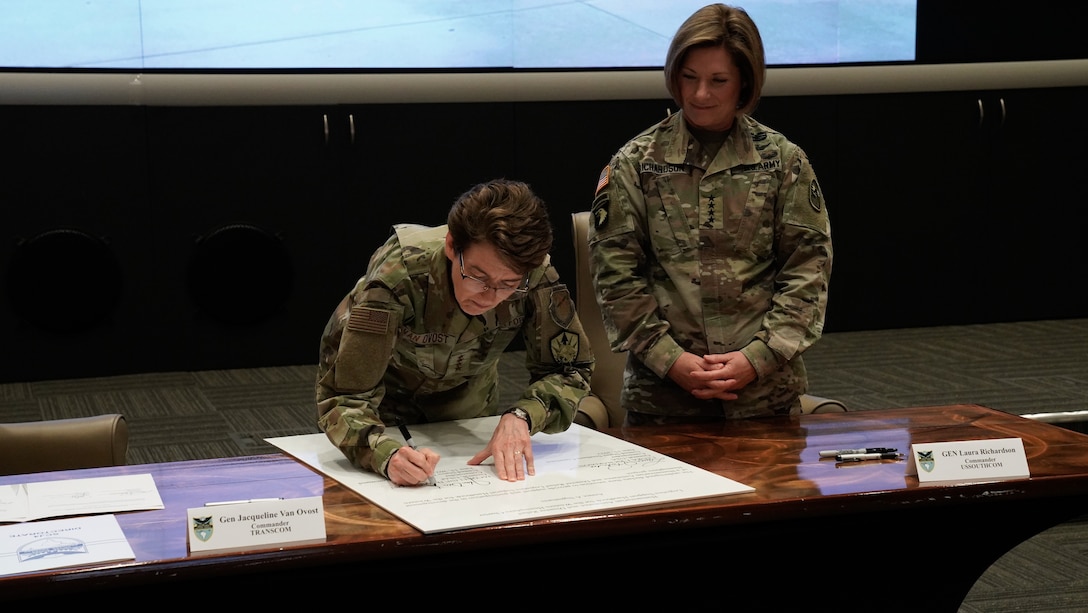 A woman signs a document as another woman looks on.
