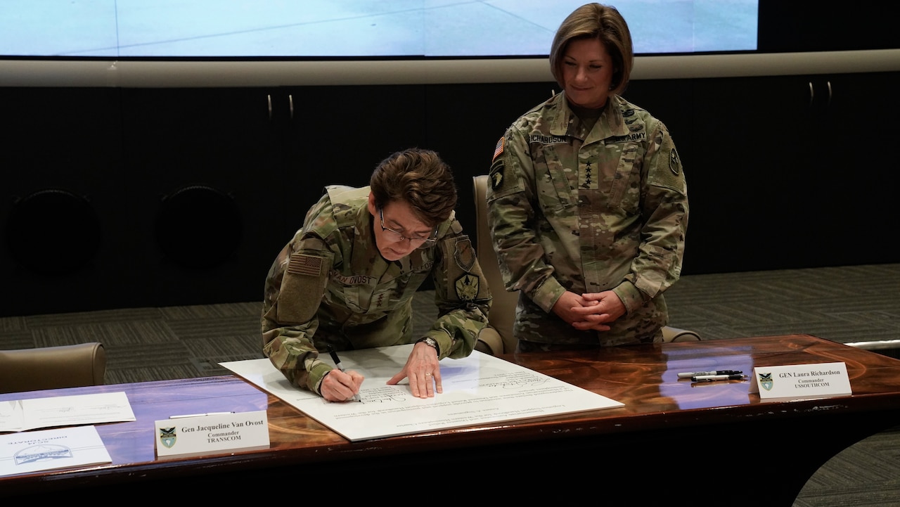 A woman signs a document as another woman looks on.