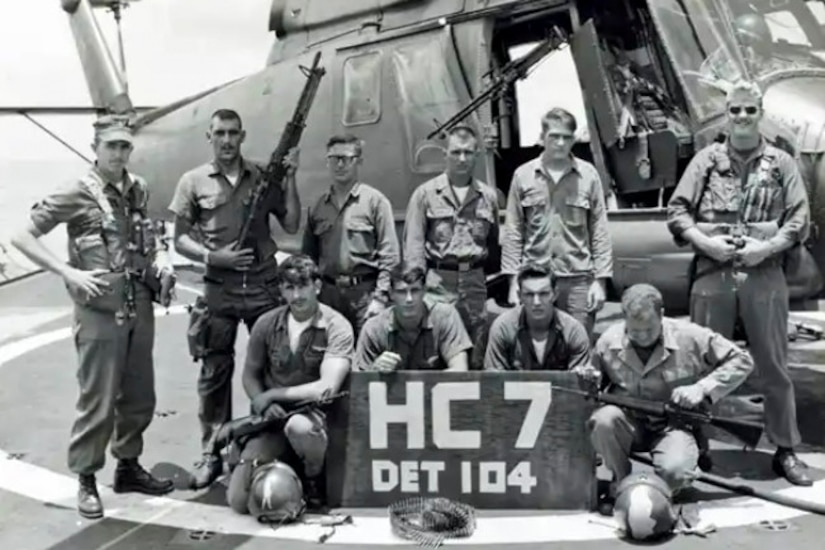 Ten men in combat dress pose for a photo in front of a helicopter and behind a sign.