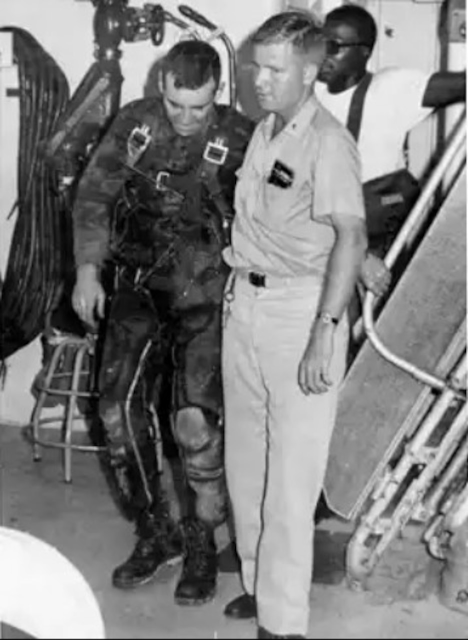 One man helps another man dressed in flight gear across a room while another person stands in the background.