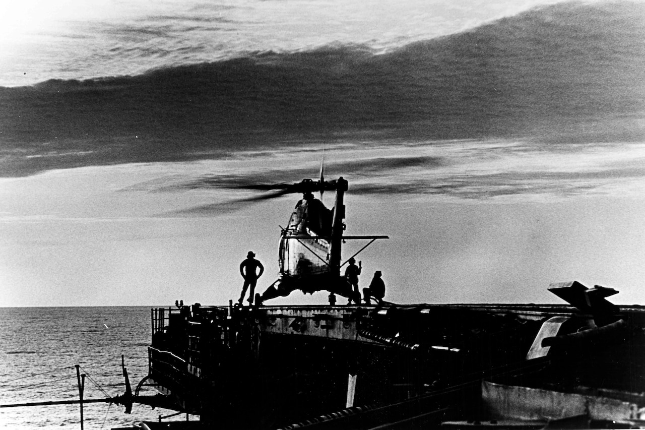 Three people stand around a helicopter that is sitting on an aircraft carrier’s flight deck.