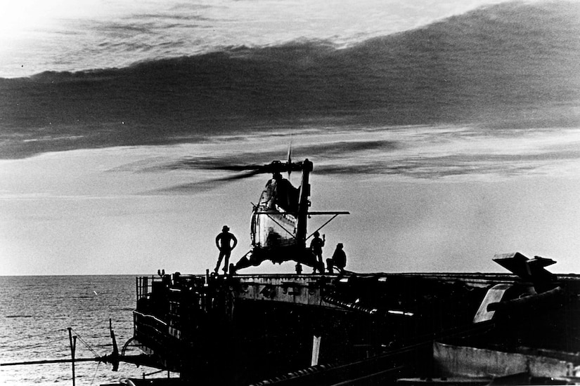 Three people stand around a helicopter that is sitting on an aircraft carrier’s flight deck.