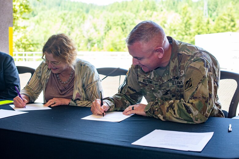 Three people sitting in an open garage bay at a table covered with a black tablecloth that reads "U.S. Army Corps of Engineers Portland District" sign a paper document.