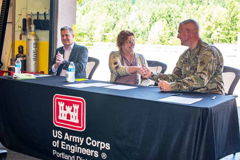 Three people sit in an open garage bay at a table covered with a black tablecloth that reads "U.S. Army Corps of Engineers Portland District." Two of the people are smiling and shaking hands.