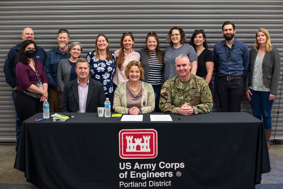 A bunch of smiling people stand in a garage bay behind a table covered with a black tablecloth that reads "U.S. Army Corps of Engineers Portland District. Three people are seated at the table in the foreground, also smiling. A paper document lies on the table in front of them.
