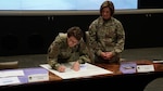 A woman signs a document as another woman looks on.