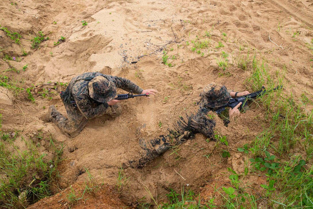 A Marine kneels beside another Marine who is covered partially in sand.