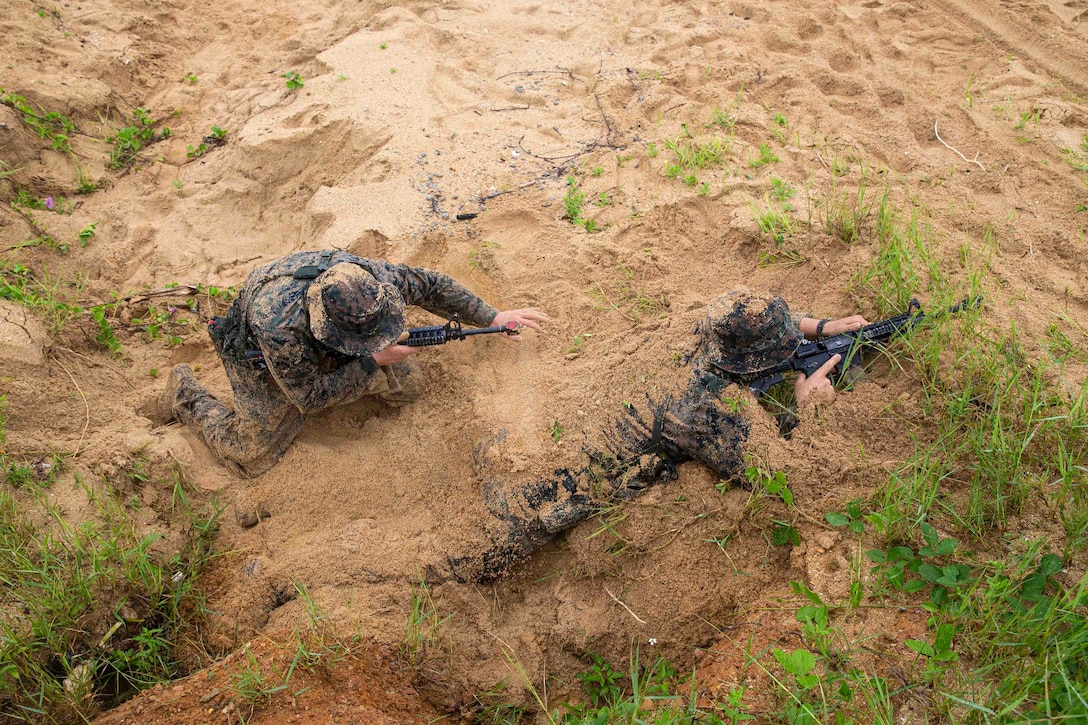 A Marine kneels beside another Marine who is covered partially in sand.