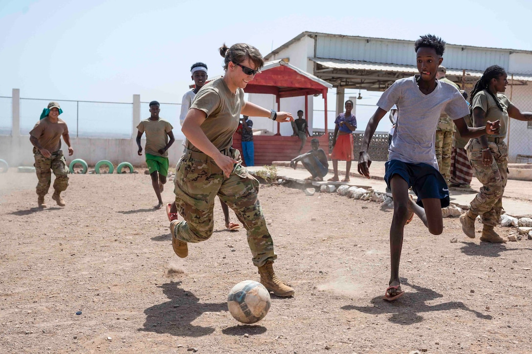 A service member runs toward a soccer ball as other run around.