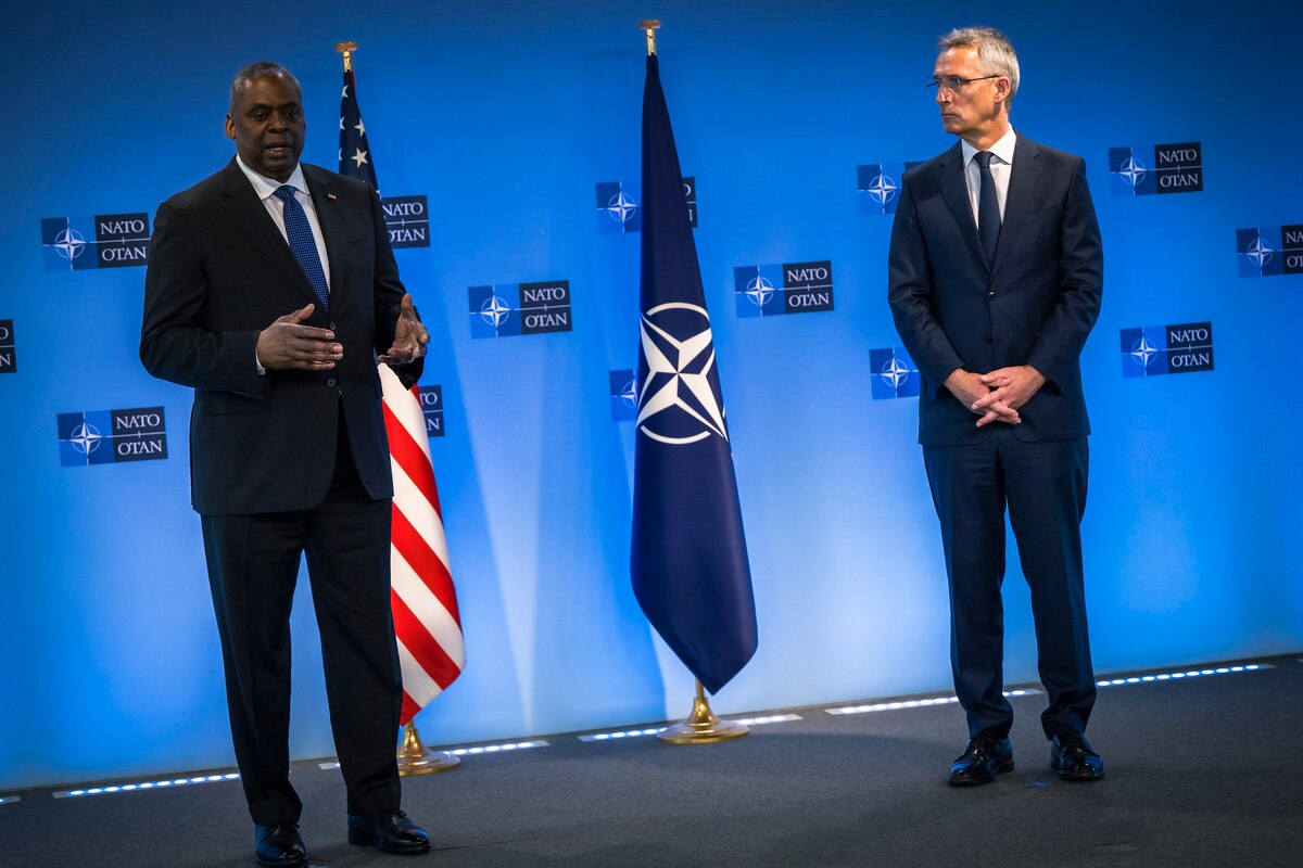 Two men wearing business suits stand on a stage with two flags in the background.