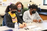 NSWC IHD Chief Technology Officer Dr. Kerry Clark sits between two students to help them figure out the best way to build their balloon buggies during their science class.